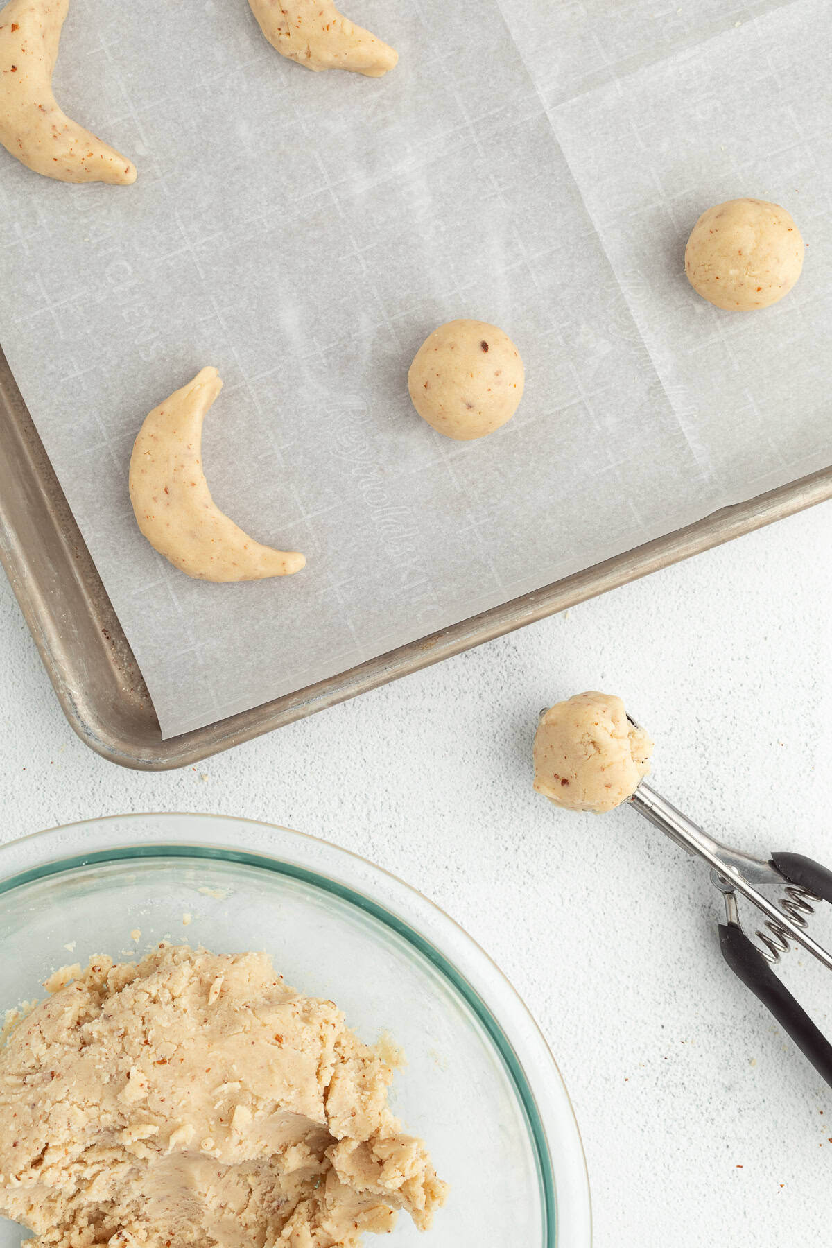 These classic almond crescent cookies have a buttery, crumbly shortbread texture and a sweet powdered sugar coating. No chilling needed! Overhead view of partially portioned and shaped almond crescent dough on a parchment-lined baking sheet next to a cookie scoop and a bowl of cookie dough.
