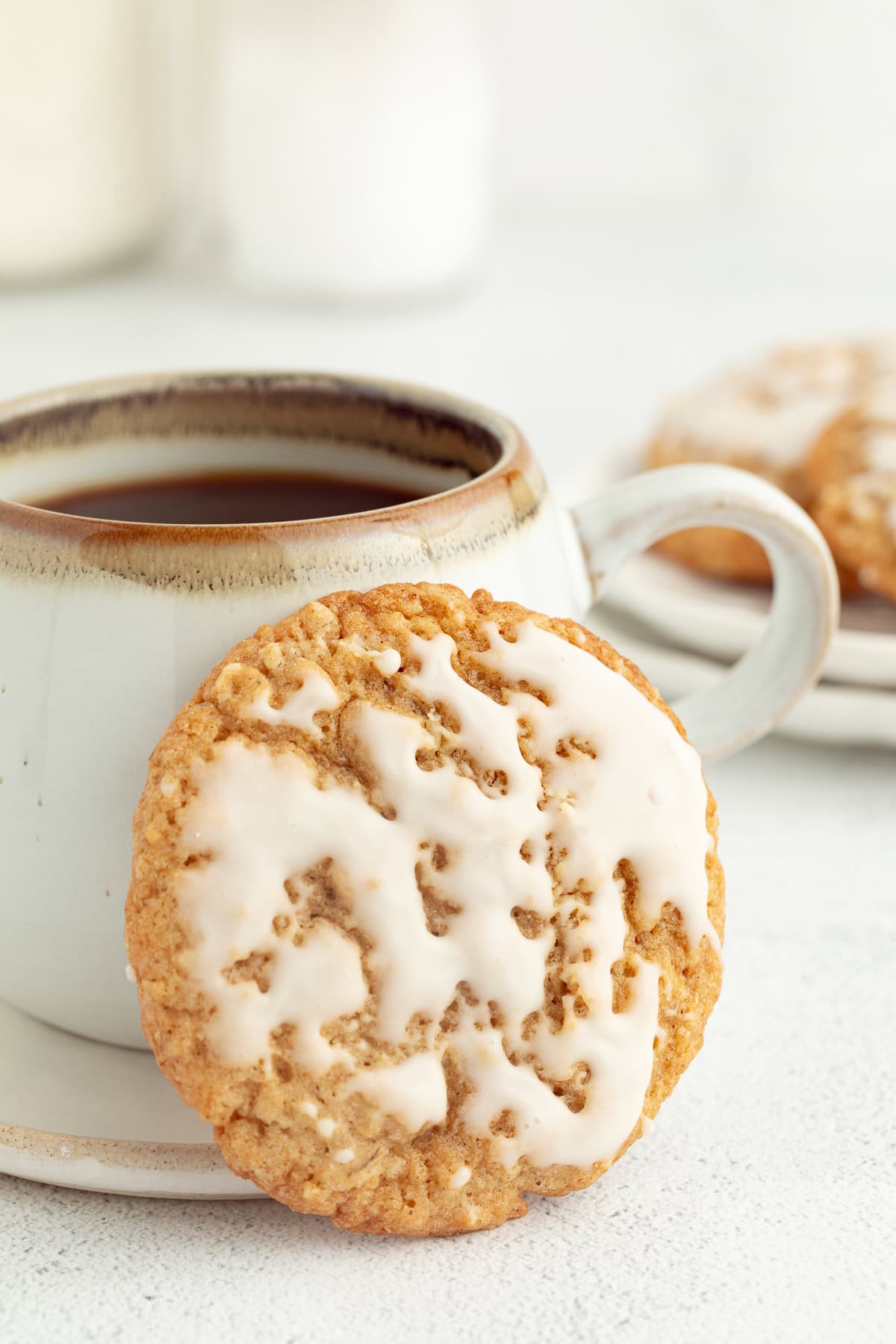 These old-fashioned iced oatmeal cookies have soft middles, crispy edges, and plenty of brown sugar and cinnamon, topped with vanilla icing. An iced oatmeal cookie propped up against a mug of coffee.