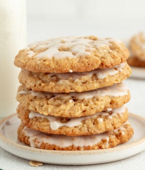 Iced oatmeal cookies stacked on a stoneware plate with a jug of milk in the background.