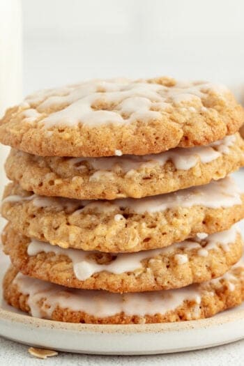 Iced oatmeal cookies stacked on a stoneware plate with a jug of milk in the background.