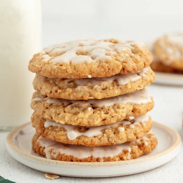 Iced oatmeal cookies stacked on a stoneware plate with a jug of milk in the background.