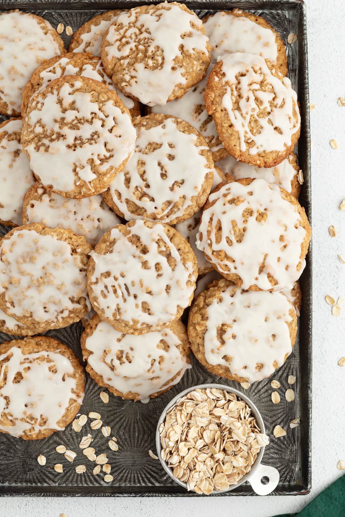 These old-fashioned iced oatmeal cookies have soft middles, crispy edges, and plenty of brown sugar and cinnamon, topped with vanilla icing. Overhead view of iced oatmeal cookies piled on a tray with a small bowl of rolled oats.