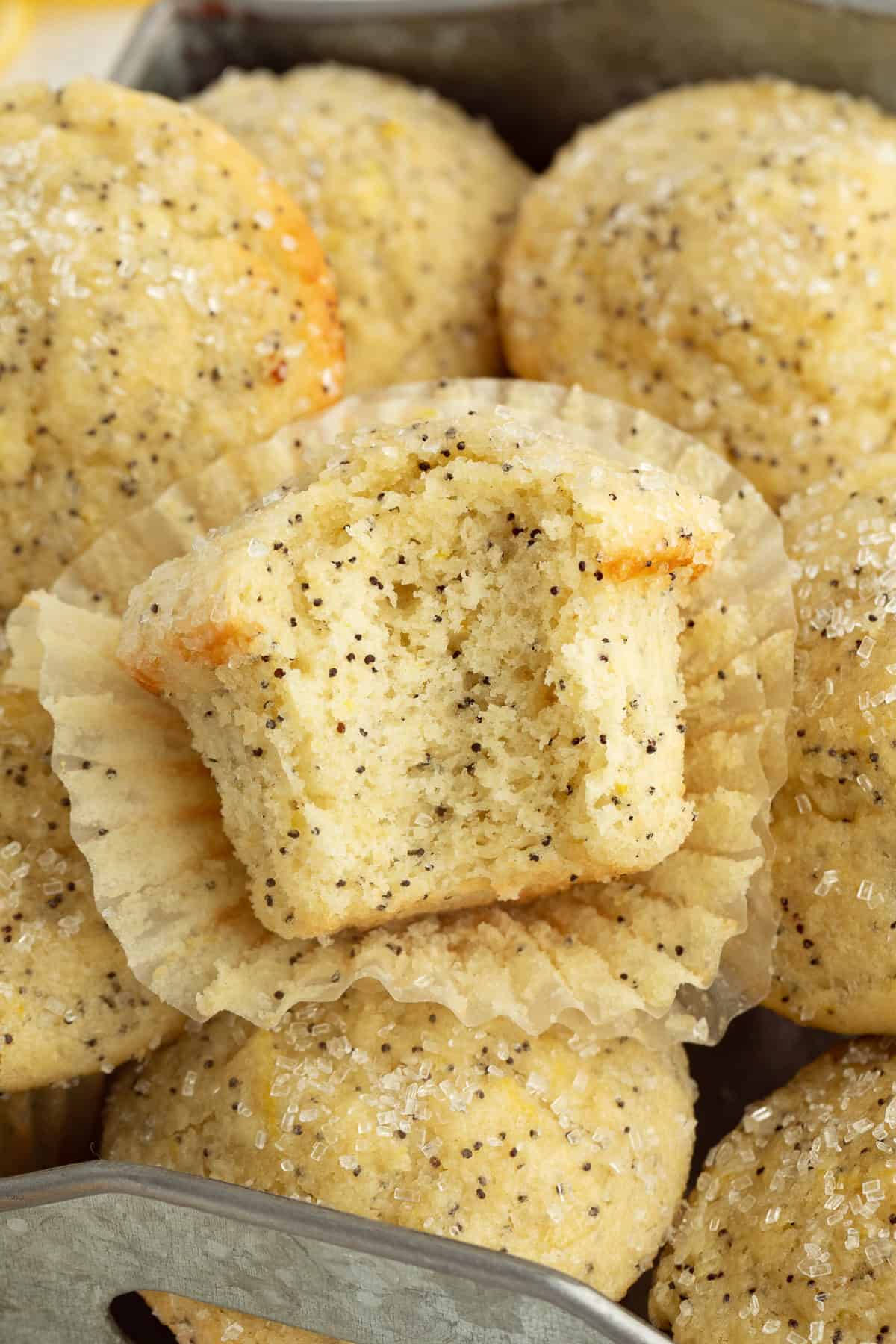 Moist, tangy, and topped with sanding sugar, these bakery-style lemon poppyseed muffins are the perfect breakfast treat. And so easy to make! A lemon poppy seed muffin with a bite missing resting on top of a pile of muffins.