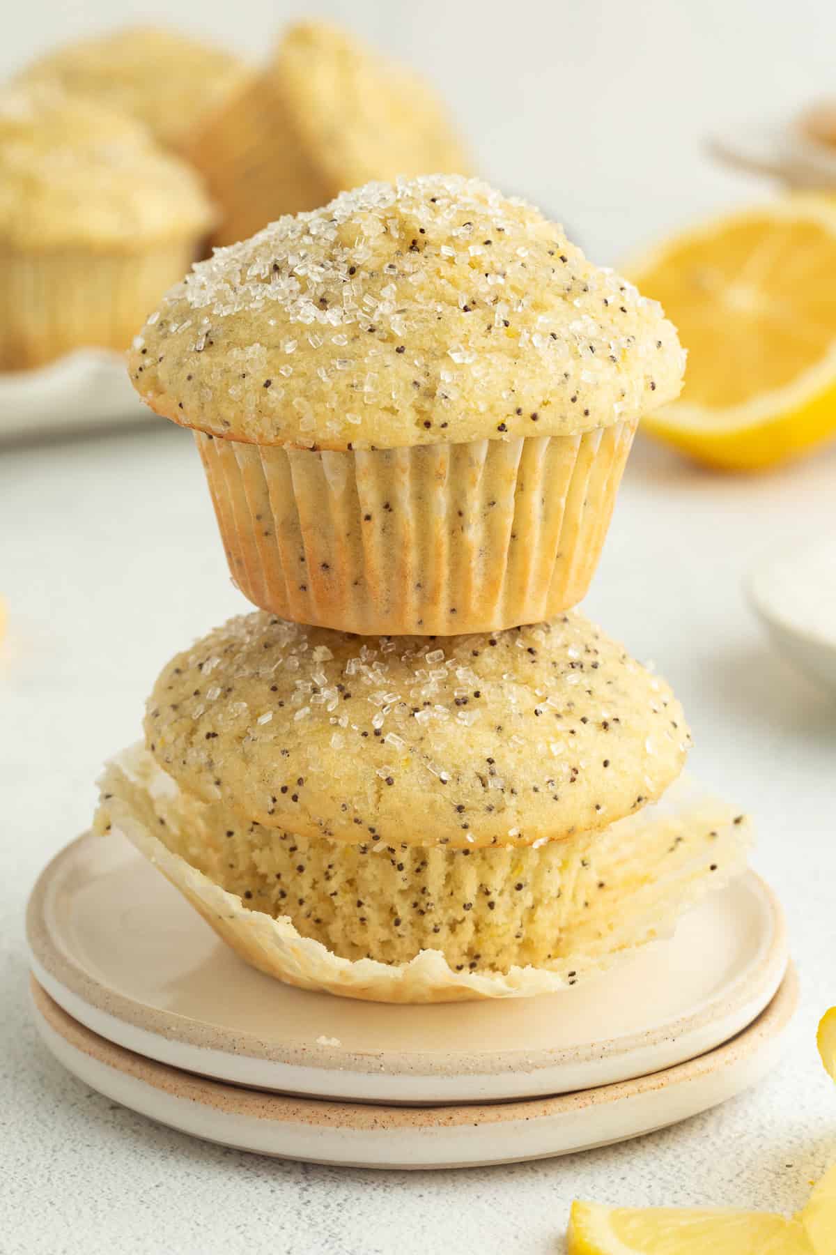 Moist, tangy, and topped with sanding sugar, these bakery-style lemon poppyseed muffins are the perfect breakfast treat. And so easy to make! Two lemon poppy seed muffins stacked on a plate with more muffins in the background.