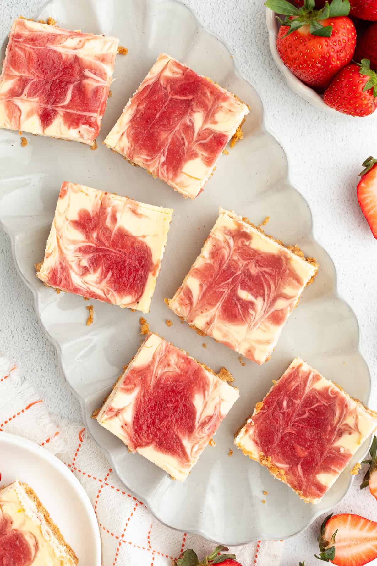 Strawberry cheesecake bars are easier to bake than a classic cheesecake, with a buttery crust and rich filling swirled with strawberry sauce! Overhead view of assorted strawberry cheesecake bars on a scalloped platter.