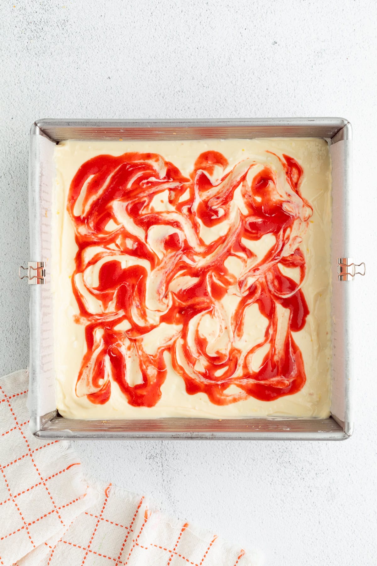 Strawberry cheesecake bars are easier to bake than a classic cheesecake, with a buttery crust and rich filling swirled with strawberry sauce! Cheesecake batter swirled with strawberry sauce in a lined square baking pan.