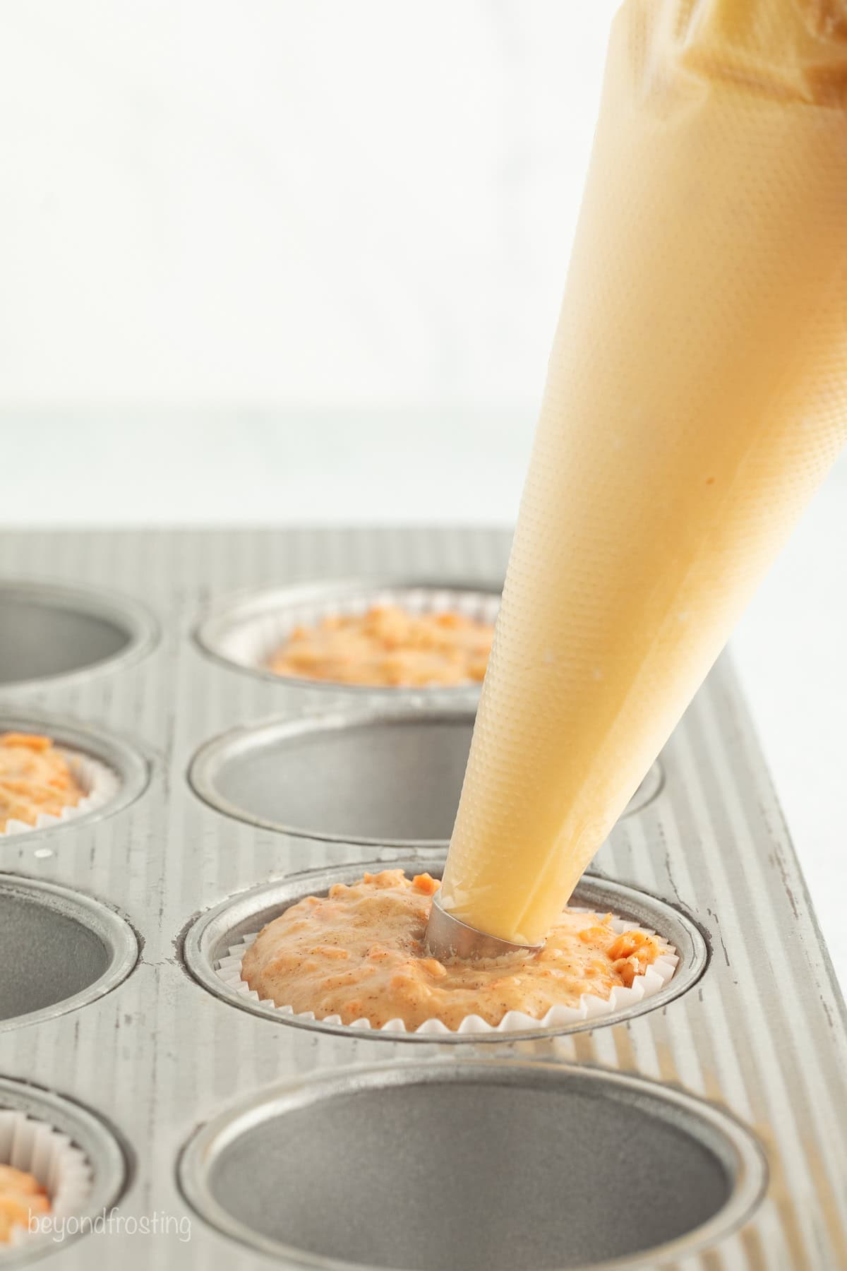 Sweet, moist carrot cake muffins are full of carrot cake flavor! They're easy to make, with the option to add a sweet cream cheese filling. A piping bag filling the center of carrot cake batter in a muffin well.