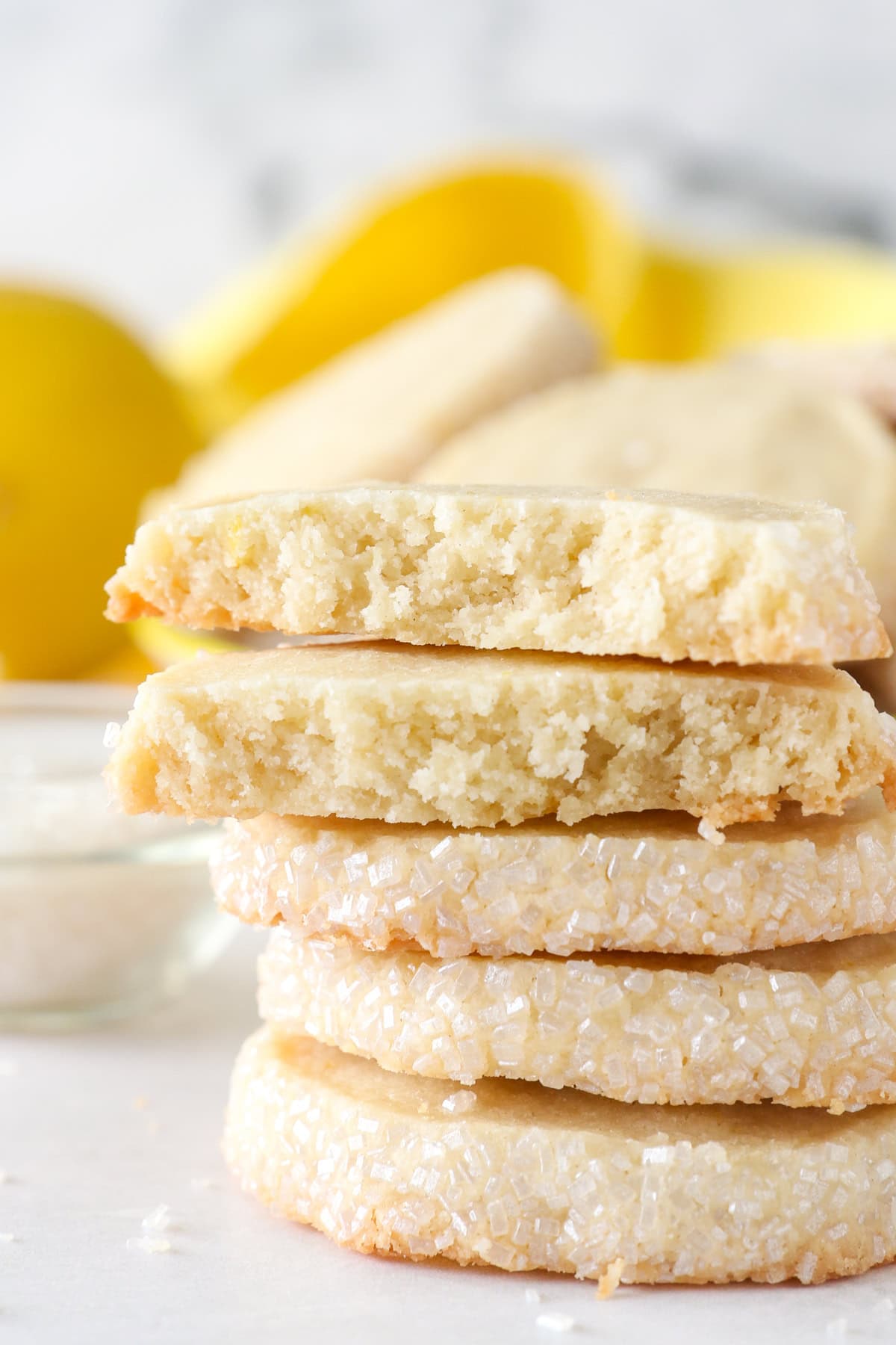These slice-and-bake lemon shortbread cookies are crisp, buttery, and melt-in-your-mouth, rolled in sanding sugar for sweet, crunchy edges. Close up of a stack of lemon shortbread cookies with the top cookie broken in half to reveal the buttery center.