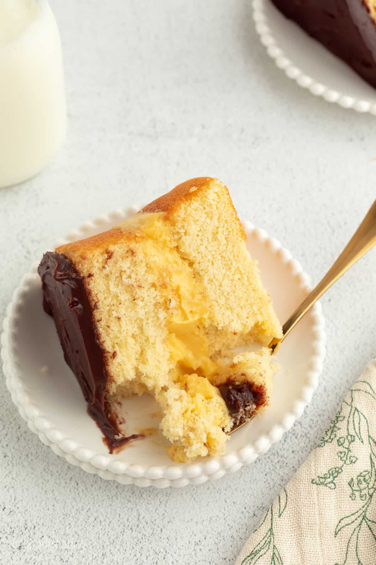This homemade Boston cream pie is an airy sponge cake filled with pastry cxream and glazed with rich chocolate ganache, all made from scratch! Overhead view of a slice of Boston cream pie with custard filling with a forkful missing.