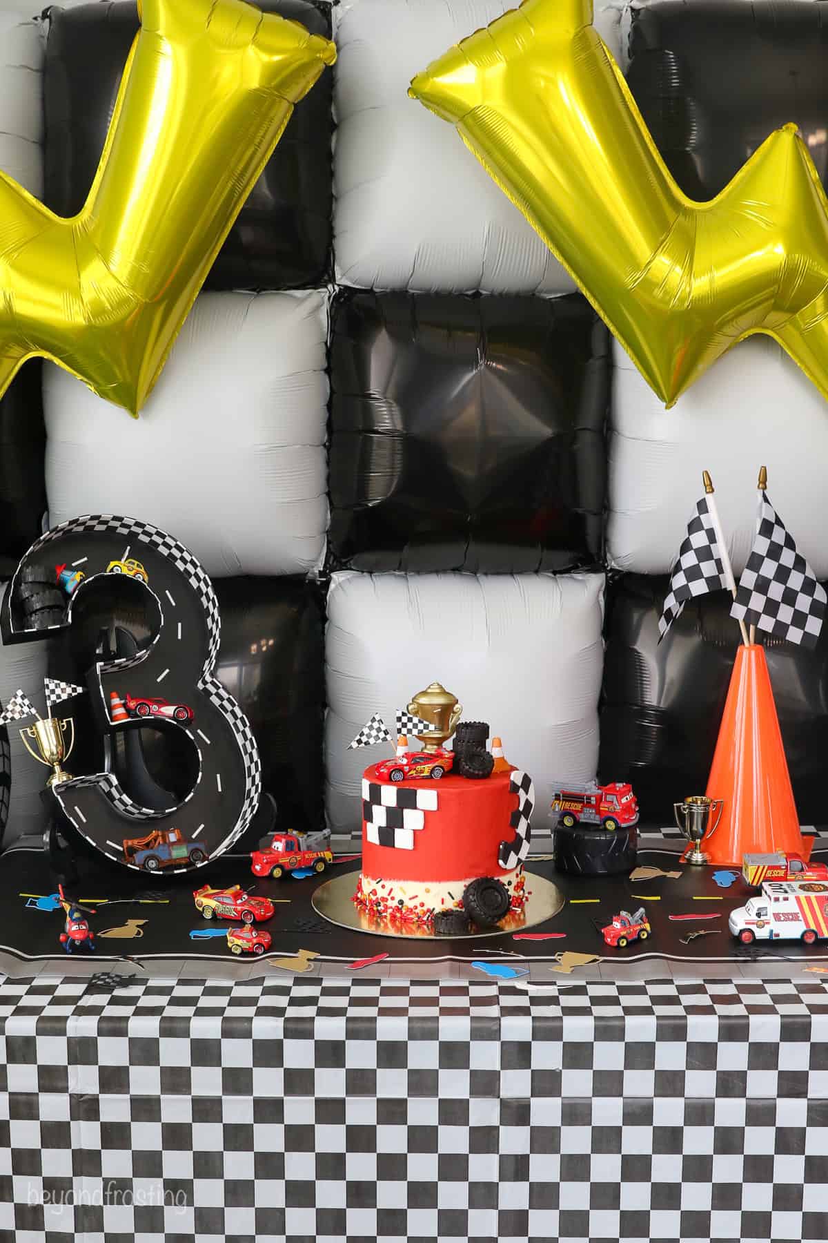 With racing-red frosting and just a little fondant, this Lightning McQueen layer cake is perfect for a kids’ Cars birthday party! A Lightning McQueen cake on a table surrounded by racing themed party decorations.