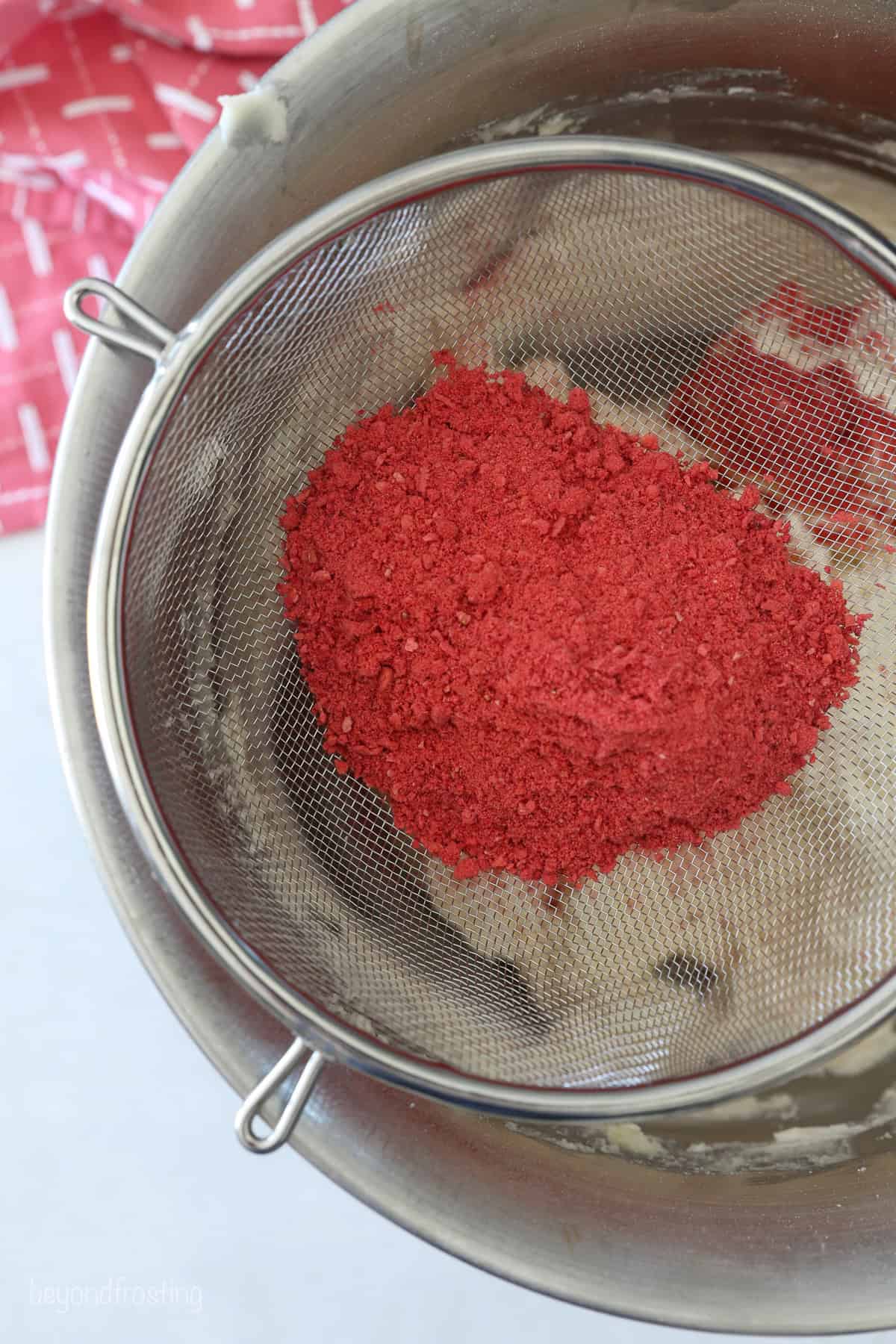 This strawberry crunch cake is a moist vanilla cake topped with creamy strawberry frosting and a crunchy strawberry and Golden Oreo crumb. Sifting freeze-dried strawberry powder into a bowl of vanilla frosting.