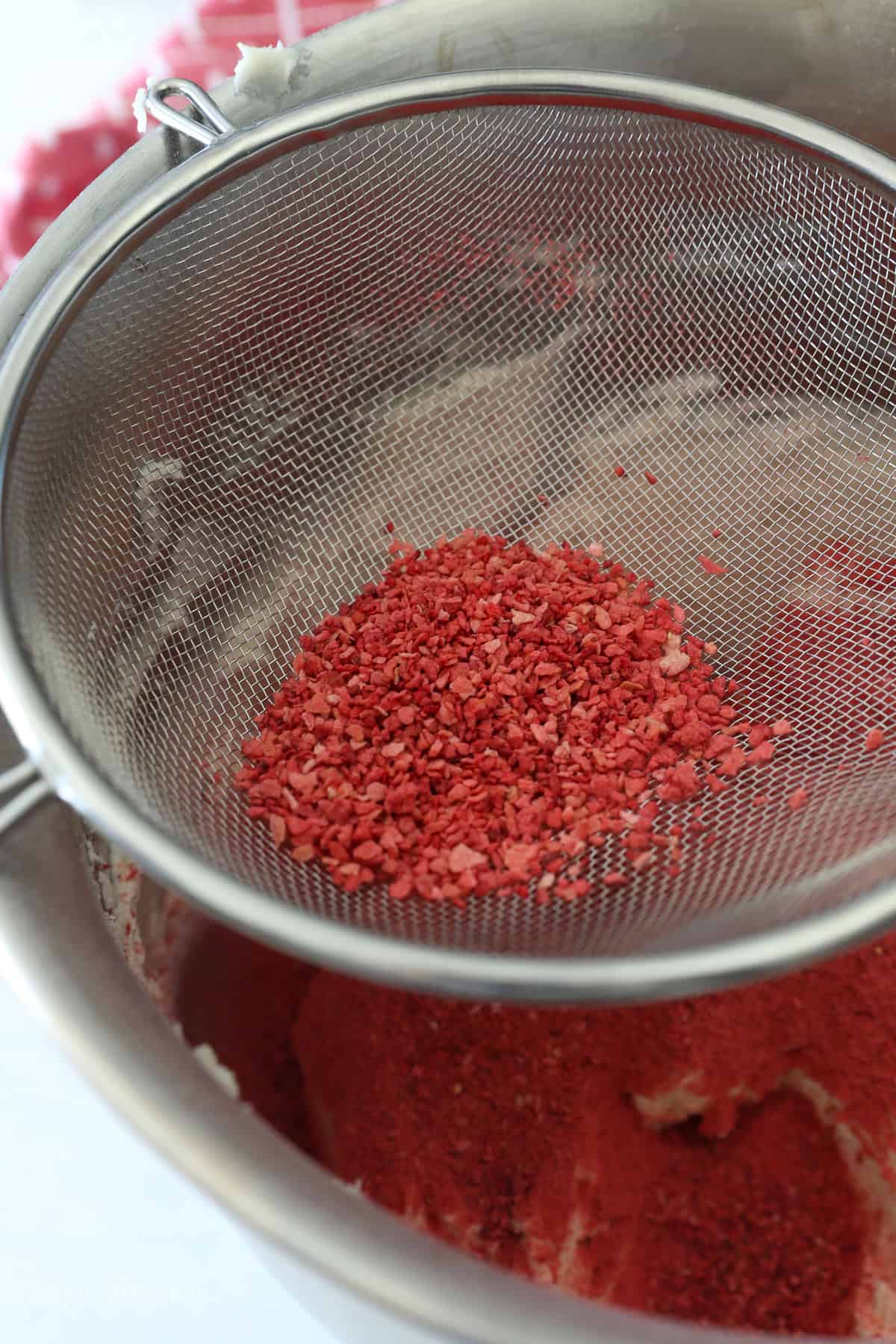 This strawberry crunch cake is a moist vanilla cake topped with creamy strawberry frosting and a crunchy strawberry and Golden Oreo crumb. Sifting freeze-dried strawberry powder into a bowl of vanilla frosting.