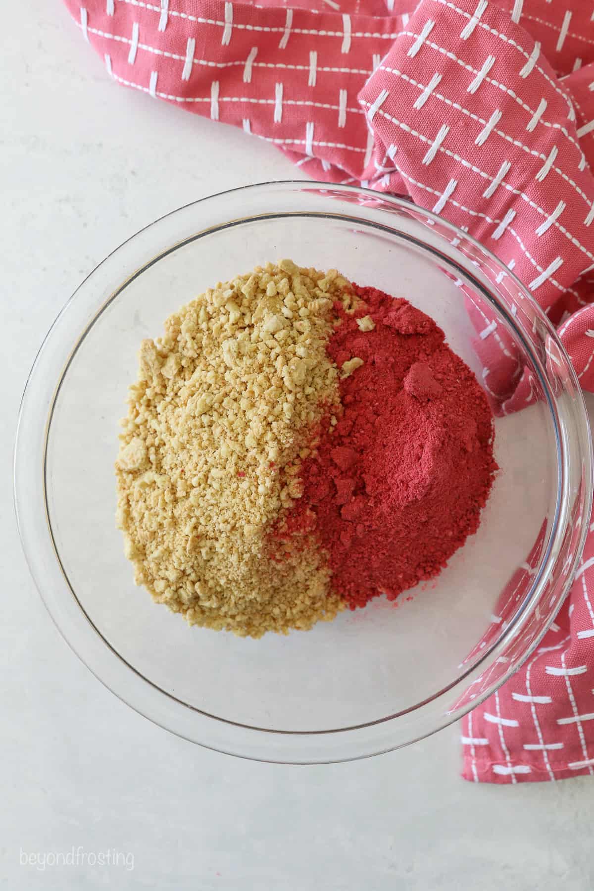 This strawberry crunch cake is a moist vanilla cake topped with creamy strawberry frosting and a crunchy strawberry and Golden Oreo crumb. Freeze-dried strawberry powder and crushed Golden Oreo crumbs added to a bowl.