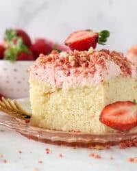 A slice of strawberry crunch cake with strawberry frosting, topped with a fresh strawberry on a plate.