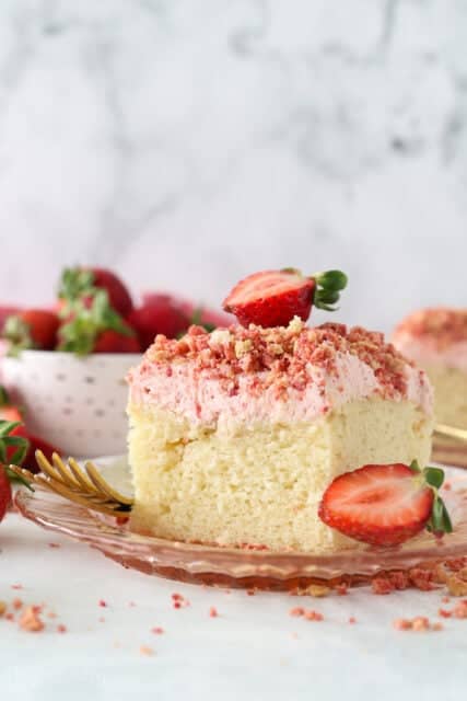 A slice of strawberry crunch cake with strawberry frosting, topped with a fresh strawberry on a plate.