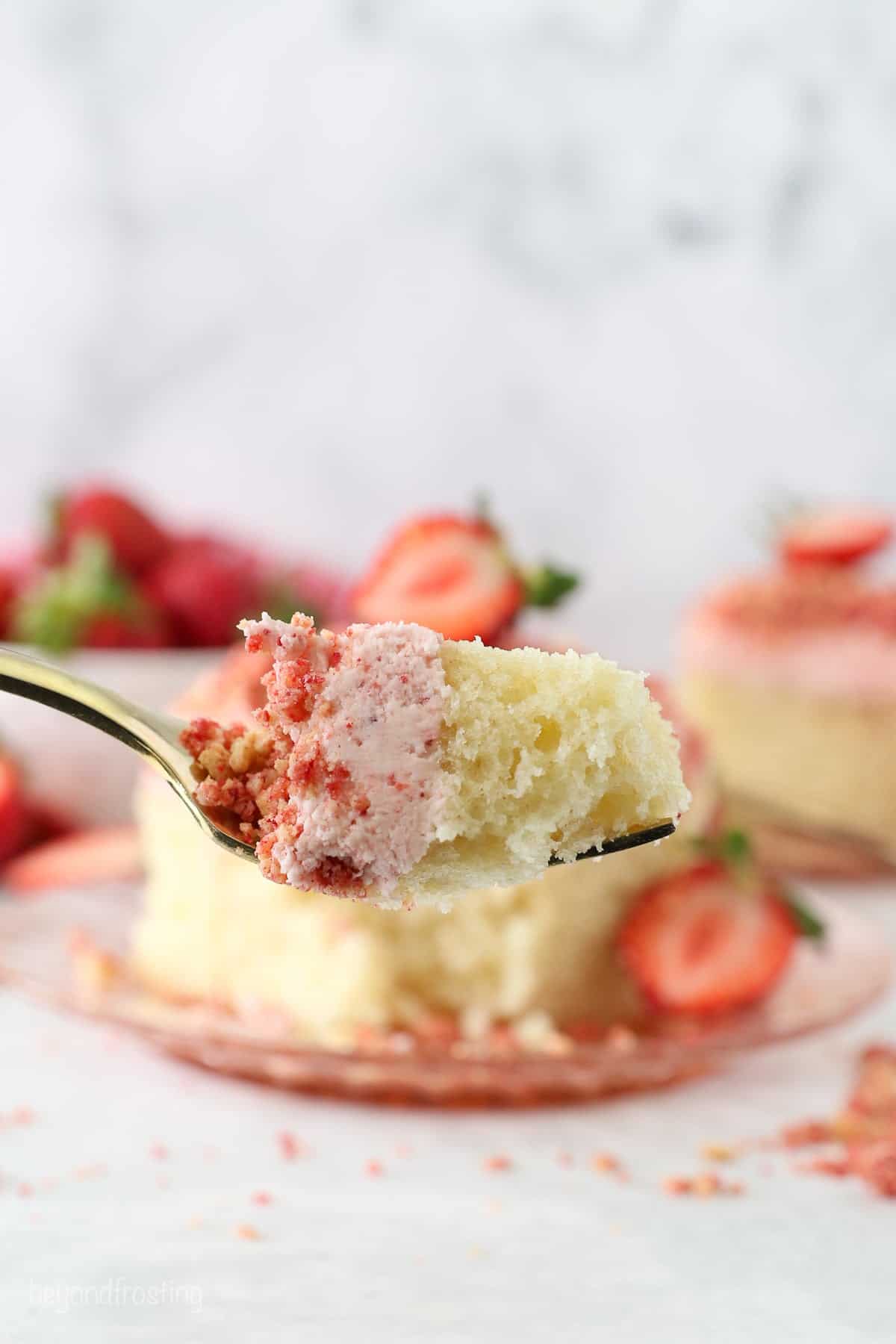 This strawberry crunch cake is a moist vanilla cake topped with creamy strawberry frosting and a crunchy strawberry and Golden Oreo crumb. Close up of a forkful of strawberry crunch cake with a cake slice in the background.