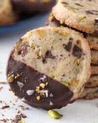 Close up of a chocolate-dipped pistachio shortbread cookie propped up against a stack of cookies.