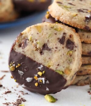 Close up of a chocolate-dipped pistachio shortbread cookie propped up against a stack of cookies.