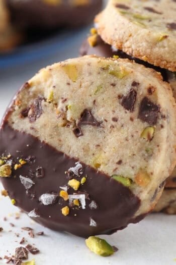 Close up of a chocolate-dipped pistachio shortbread cookie propped up against a stack of cookies.